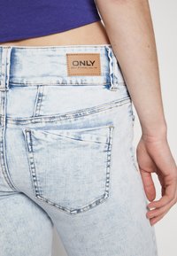 Persona vestida con jeans de mezclilla azul claro con efecto acid wash y una etiqueta de cuero marrón con la inscripción "ONLY" en la cinturilla, de pie contra un fondo blanco.
