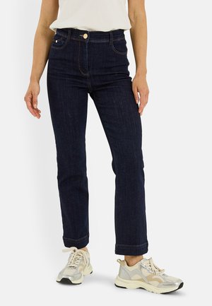 Mujer con vaqueros azul oscuro de corte recto, zapatillas blancas con detalles beige y plateados, y una camiseta blanca metida por dentro, sobre un fondo liso.