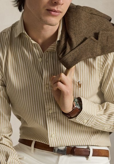 Homme en chemise beige rayée à boutons porte une veste marron à chevrons sur l'épaule, avec une montre et une ceinture en cuir marron et un pantalon blanc.