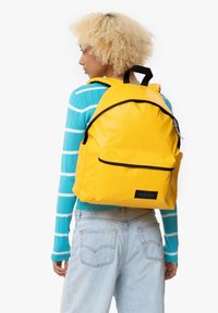 Mochila amarilla con detalles en negro, fabricada con material duradero. Cuenta con un bolsillo frontal y correas ergonómicas, vista sobre un fondo liso.
