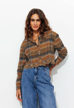 Chemise à carreaux brune, courte, avec des manches longues et une fermeture à boutons, ornée de broderies florales sur l'ourlet. Associée à un jean bleu taille haute.