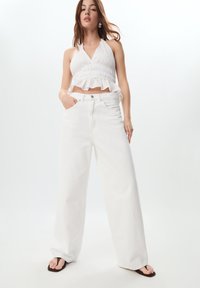 Top corto bianco con scollatura a halter e orlo arricciato abbinato a jeans bianchi a vita alta e gamba larga. Modella indossa ciabatte marroni. Sfondo chiaro.