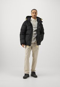 Black puffer jacket with a hood, zip closure, yellow accents, paired with a light-colored pullover and cargo pants, completed with dark footwear.