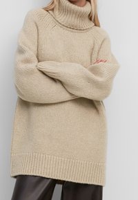 Beige neulottu pooloneule, leveillä hihoilla, resorilla helmassa ja sileällä pinnalla, ulottuu lantiota pidemmälle.