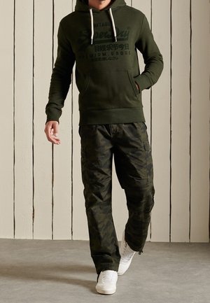 Grön hoodie med dragsko och en framficka, kombinerad med byxor i kamouflagemönster. Vita sneakers kompletterar outfiten.