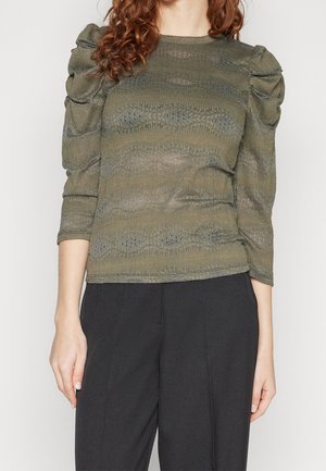 Vrouw draagt een olijfgroen gebreid topje met pofdrie-kwart mouwen en zwarte wijde broekspijpen, staand tegen een effen witte achtergrond.