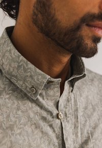 Lichtgrijs shirt met een bloemenpatroon, voorzien van een button-down kraag en subtiele texturen. De knopen zijn wit en rond.