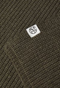 Tissu tricoté vert présentant une texture côtelée, avec une étiquette blanche affichant un logo stylisé en noir dans le coin inférieur droit.