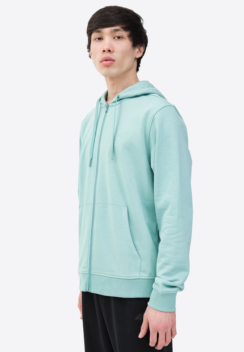 Sudadera con cremallera de color verde menta claro, con bolsillos frontales, capucha ajustable y puños acanalados. Tejido suave con un sutil detalle de textura.