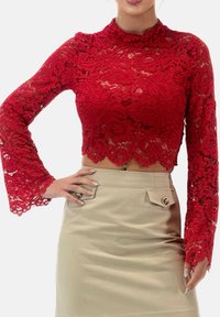 Top corto de encaje rojo con mangas largas acampanadas, con patrones florales y dobladillo con bordes ondulados. Combina con una falda beige con detalles de botones.