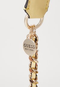 Kultaketju, jossa on keltainen tekonahkainen hihna, sisältää pyöreän hopeisen charm-palan, johon on kaiverrettu "GUESS EST.1981." Liukas pinta ja kiiltävät yksityiskohdat.