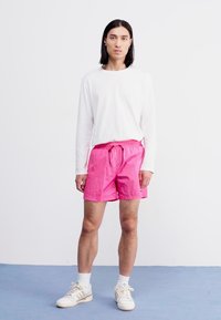 Lyserøde svømmedragter med elastisk talje og justerbar snor, pairret med en hvid langærmet skjorte og lyse sneakers.