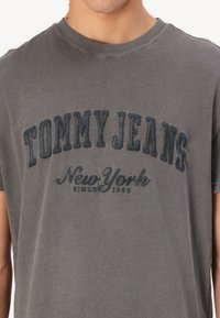 Camiseta de algodón gris con un logotipo bordado texturizado en azul marino que dice "TOMMY JEANS New York desde 1985". Cuello redondo y mangas cortas.