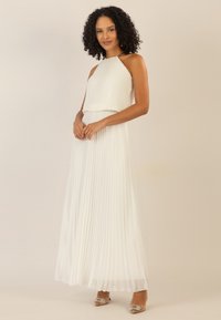 Weißes, plissiertes Maxikleid mit Halterneck und geschichteter Brustpartie. Aus transparentem Material und mit fließender Silhouette. Akzentuiert mit glitzernden Absätzen.