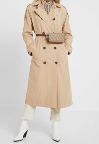 Béžový dlouhý trenčkot s dvouřadými knoflíky, nosený přes vzorovaný top, spárován s bílými podpatkovými botami a béžovou ledvinkou.