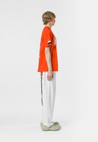 Camiseta roja de gran tamaño con un estampado gráfico, combinada con pantalones blancos y zapatos verdes texturizados; la vista lateral muestra un diseño de outfit casual.