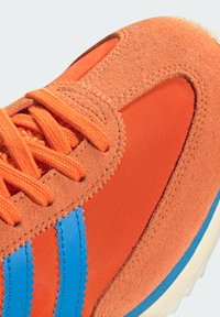 Oranje sneaker van een combinatie van suede en synthetisch materiaal, met blauwe strepen, gestructureerde overlays en oranje veters.