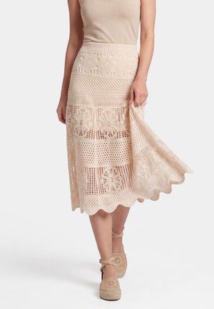 Frau trägt einen beigen, gehäkelten Midirock mit Blumenmuster, hält den Rocksaum und kombiniert diesen mit beigen Espadrilles mit Knöchelriemen.