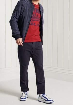 Chaqueta bomber de la marina, camiseta gráfica roja, pantalones oscuros y zapatillas azul/blancas. Incluye características como tejido suave y corte relajado.