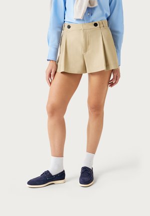 Donna che indossa pantaloncini beige a pieghe con bottoni neri, camicia a maniche lunghe azzurro chiaro, calzini bianchi e scarpe stringate blu navy.
