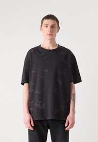 Svart oversized T-skjorte med et teksturert, litt falmet design. Har rund hals og korte ermer. Komplementeres av mørke jeans.