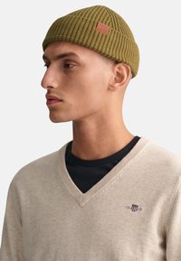 Olivegrüner Strickmütze mit geripptem Muster, versehen mit einem Lederetikett. Getragen mit einem beigen V-Neck-Pullover über einem schwarzen Shirt.