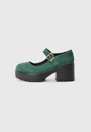 Pantof verde din suede tip Mary Jane, cu un toc gros din platformă neagră și un curea cu cataramă. Afișat pe un fundal alb simplu.