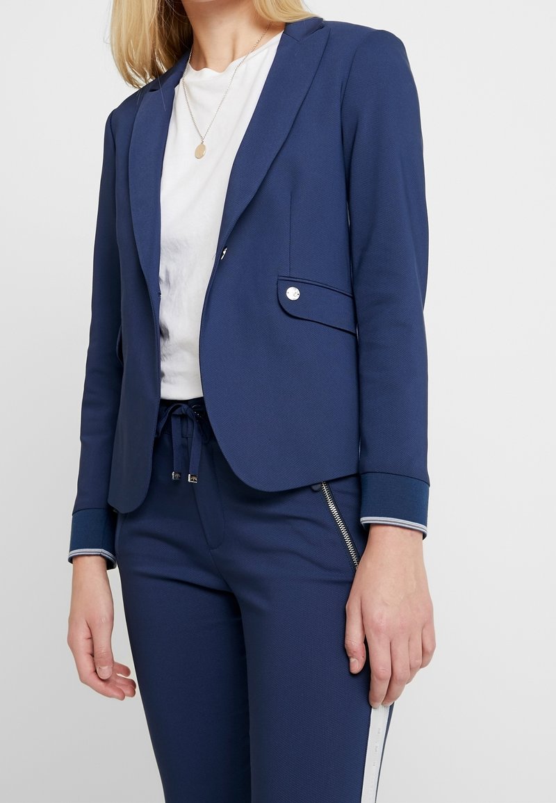Femme portant un blazer bleu marine et un pantalon assorti, avec une chemise blanche et un collier pendentif sur un fond clair uni.