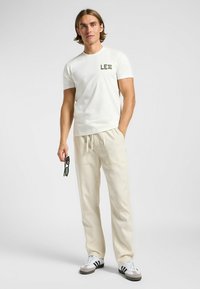 Camiseta blanca con logo verde "LE", combinada con pantalones beige con cordón y gafas de sol negras. El calzado presenta franjas en blanco y negro.