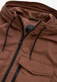 Veste à capuche marron avec cordons noirs, fermeture éclair centrale noire et une poche à rabat sur la poitrine.