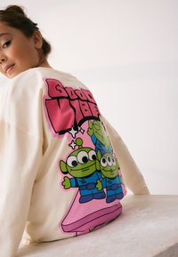 Sweatshirt in cream, featuring a large colorful graphic of cartoon aliens on the back, with "Good Vibes" text in bold pink.