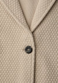 Blazer en tissu beige texturé avec un col cranté et un bouton rond noir gravé de texte. Motif subtil à travers le matériau.