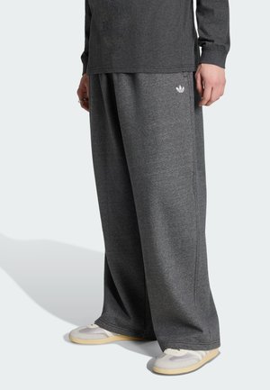 Persona che indossa pantaloni della tuta grigio scuro a gamba larga con logo Adidas bianco, maglietta a maniche lunghe coordinata e sneakers bianche con suola beige.