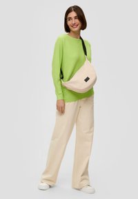 Helle rosa Umhängetasche mit glatter Textur, schwarzem Riemen und einem dezenten Etikett. Kombiniert mit einem leuchtend grünen gerippten Pullover und beigen Hosen.