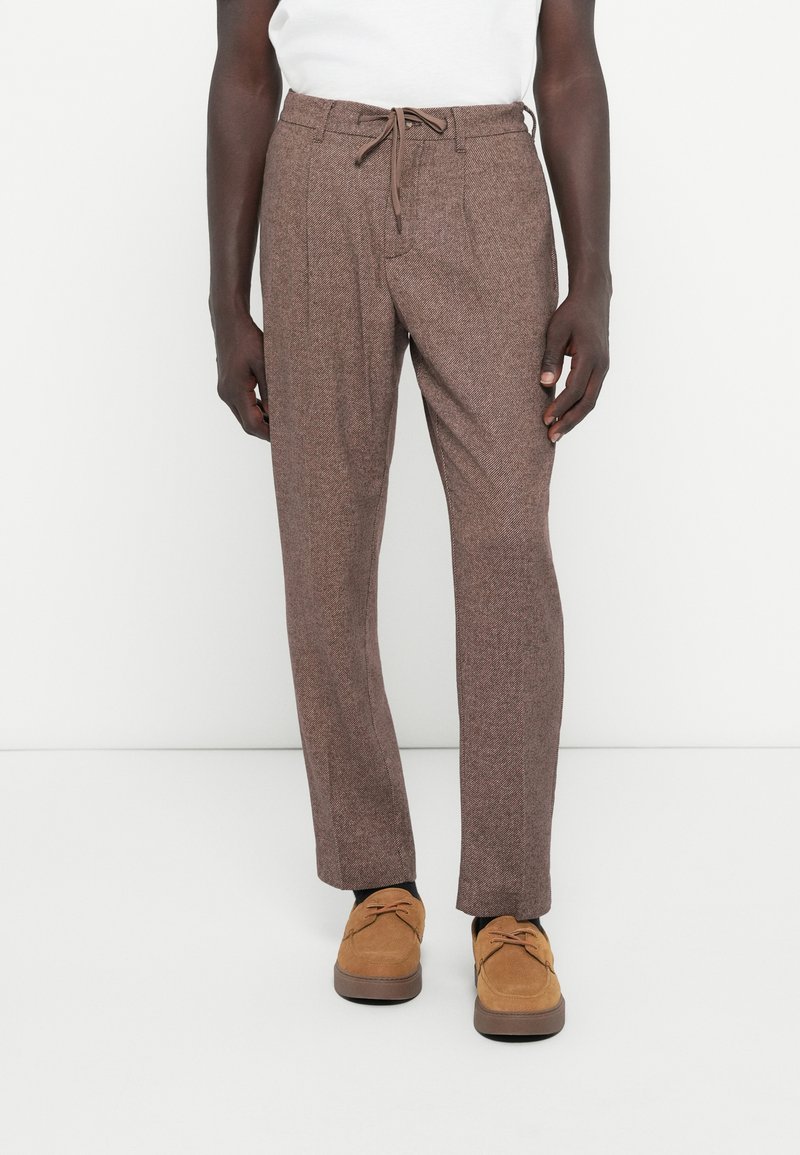 Brown textured trousers with a drawstring waist, featuring side pleats and straight legs, paired with brown suede shoes.