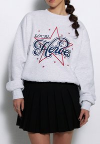 Szary sweatshirt z grafiką "LOCAL HEROES" w niebieskim i czerwonym kolorze, z motywem gwiazdy oraz gwiazdkami, w połączeniu z czarną plisowaną spódnicą.