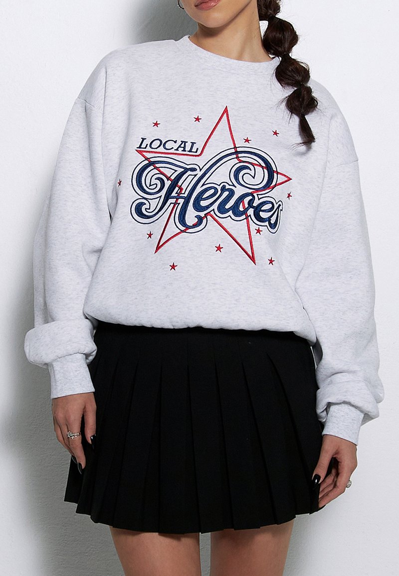 Szary sweatshirt z grafiką "LOCAL HEROES" w niebieskim i czerwonym kolorze, z motywem gwiazdy oraz gwiazdkami, w połączeniu z czarną plisowaną spódnicą.