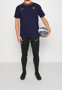 Námořnický sportovní dres s vínovým límcem a zlatým logem, spárovaný s černými slim fit kalhotami a černými kopačkami, držící šedý fotbal.