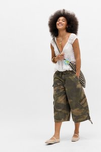 Hvit bluse med korte ermer og volanger; kamuflasje cargo-shorts; vevd svart og hvit veske med en blå blomst; beige flate sko; krøllete hår.