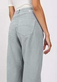 Vaaleansiniset corduroypöksyt, joissa on leveät lahkeet, takataskut, teksturoidut ribbikuvio ja korkea vyötärö. Lähikuva takanäkymästä ja käden sijoittelusta.