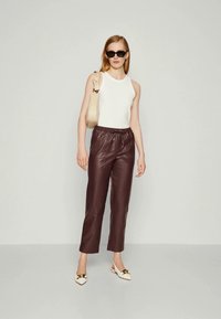 Femme portant des lunettes de soleil noires, un haut sans manches blanc, un pantalon en cuir marron foncé, un sac à bandoulière beige et des chaussures plates blanches avec des détails dorés.