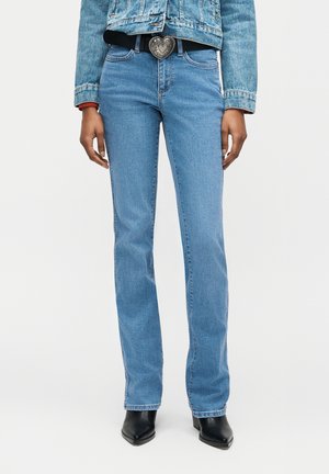 Person som har på seg lyseblå jeans med rette ben, svarte spisse støvler, denimjakke og beltespenne formet som et hjerte.