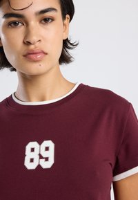 Burgundin lyhythihainen t-paita valkoisilla kuvoilla, jossa rintamuksessa suuri valkoinen '89' printti. Puuvillakangasta, jolla on sileä tekstuuri.