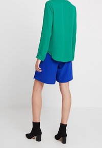 Grön blus med lång ärm och rundad nederkant, matchad med blå shorts. Svarta klackskor med blockklack fulländar outfiten.