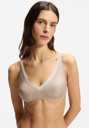 Frau mit langen braunen Haaren, die einen transparenten beigen BH mit breiten Trägern trägt, schaut in die Kamera vor einem schlichten weißen Hintergrund.