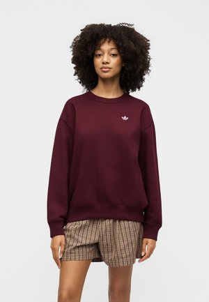 Ung kvinde med krøllet hår iført en vinrød Adidas sweatshirt og ternede shorts, stående mod en ensartet lys baggrund.