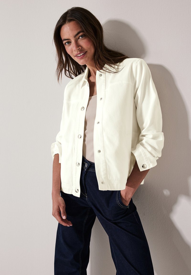 Witte button-up jas met een relaxed fit, voorzien van een platte kraag, zilveren drukknoopjes en opgerolde mouwen, gecombineerd met donkerblauwe broek.