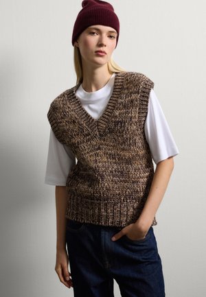 Jonge vrouw met een bordeauxrode beanie, een bruine gebreide mouwloze trui over een witte blouse en donkerblauwe jeans, staande tegen een effen muur.