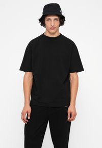 Camiseta negra de manga corta, hecha de algodón. Diseño simple, corte relajado, con un pequeño logo en el dobladillo. Combinada con pantalones negros.