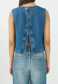 Trendi izrezani denim top, plave boje, s dizajnom na leđima koji ima prorez i kopčanje na dugme. Uparen s svijetloplavim trapericama visokog struka, koje prikazuju stražnje džepove.